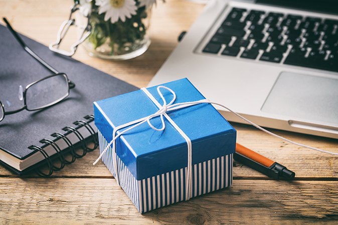 A blue striped gift box sitting on a desk with a laptop, notebook, eyeglasses and a pen